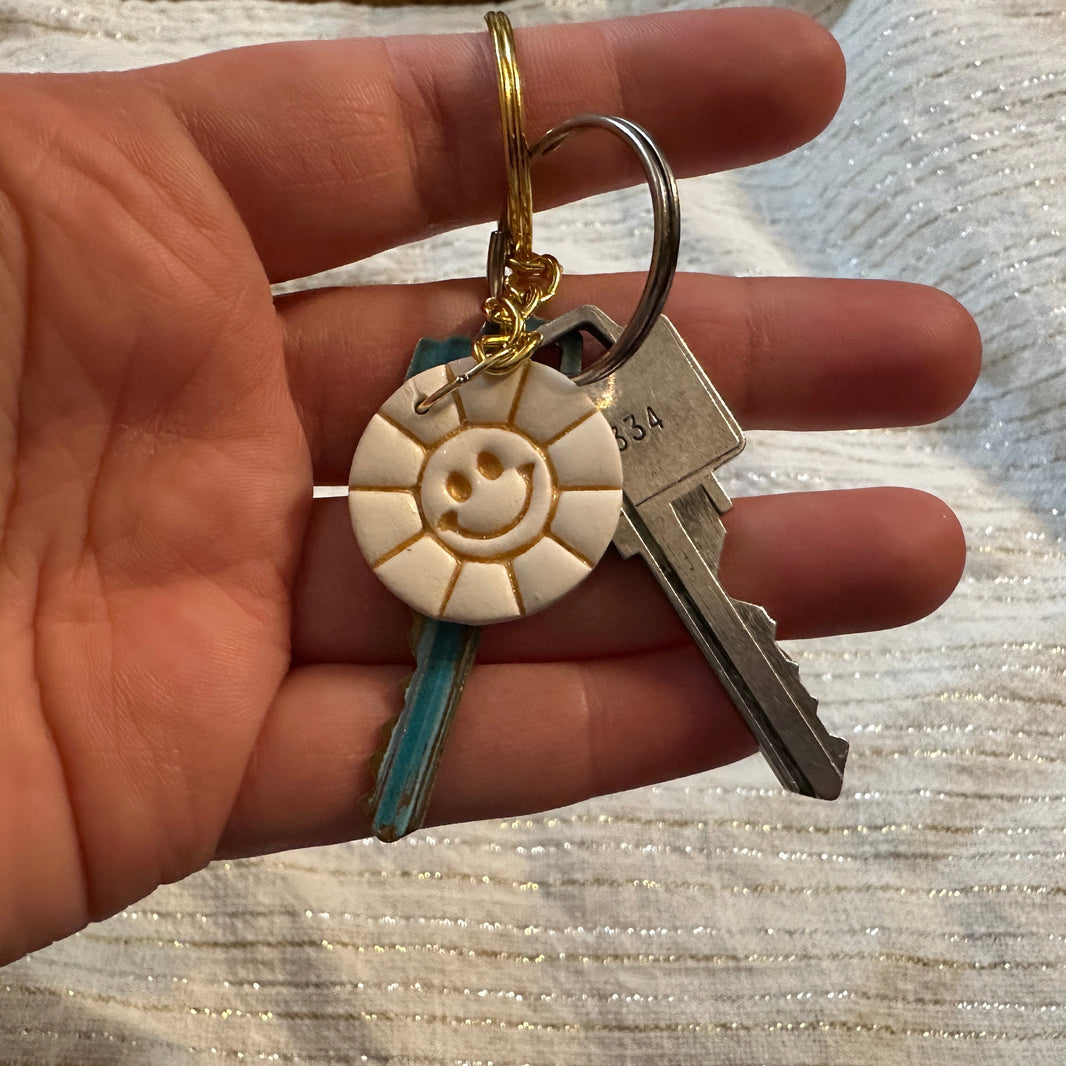 Hand holding a round, white polymer clay keychain featuring a smiley face sun design, and embossed metallic gold liquid clay details.