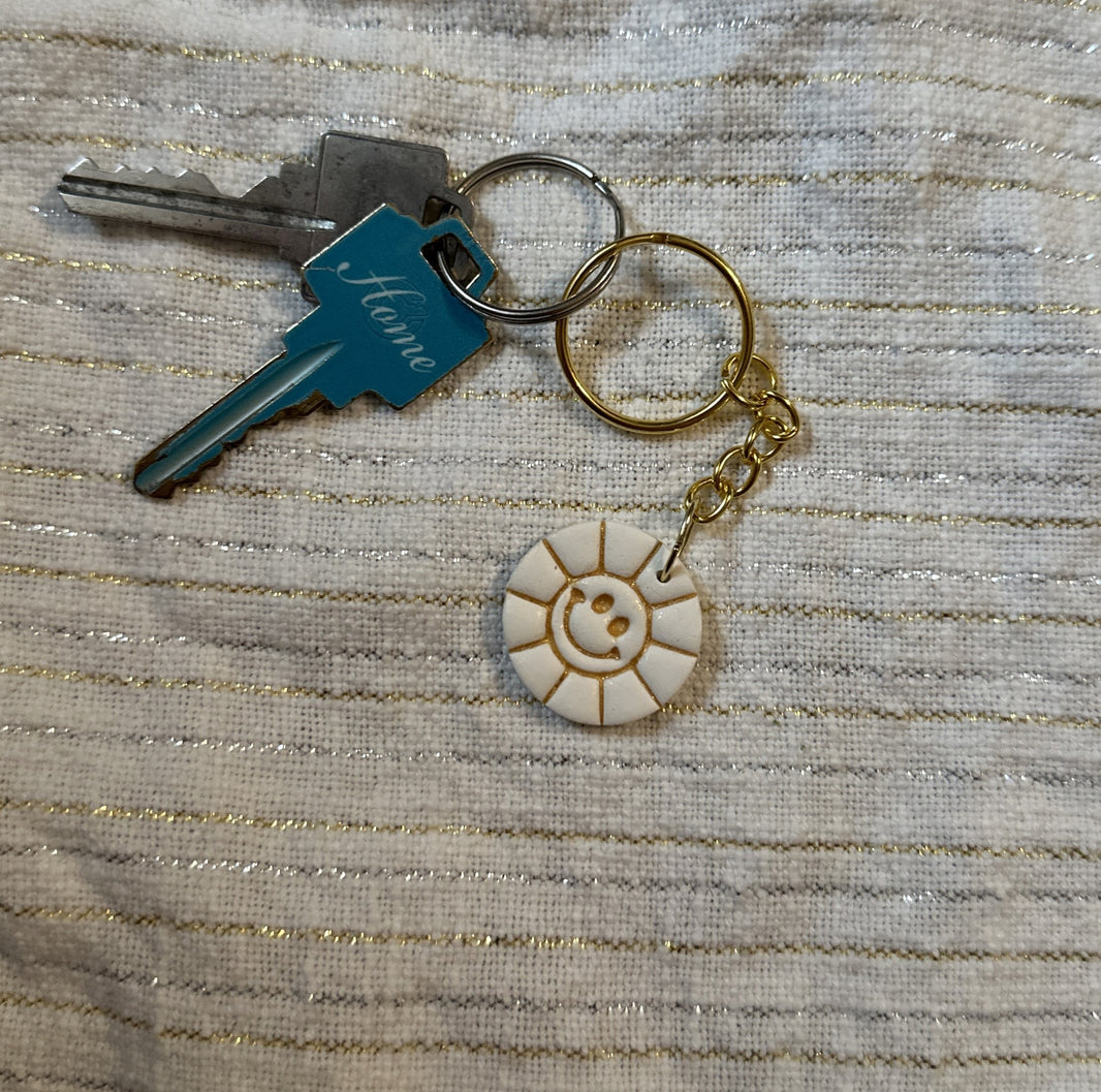 A round, white polymer clay keychain featuring a smiley face sun design, and embossed metallic gold liquid clay details, with a set of keys on a white cotton background.