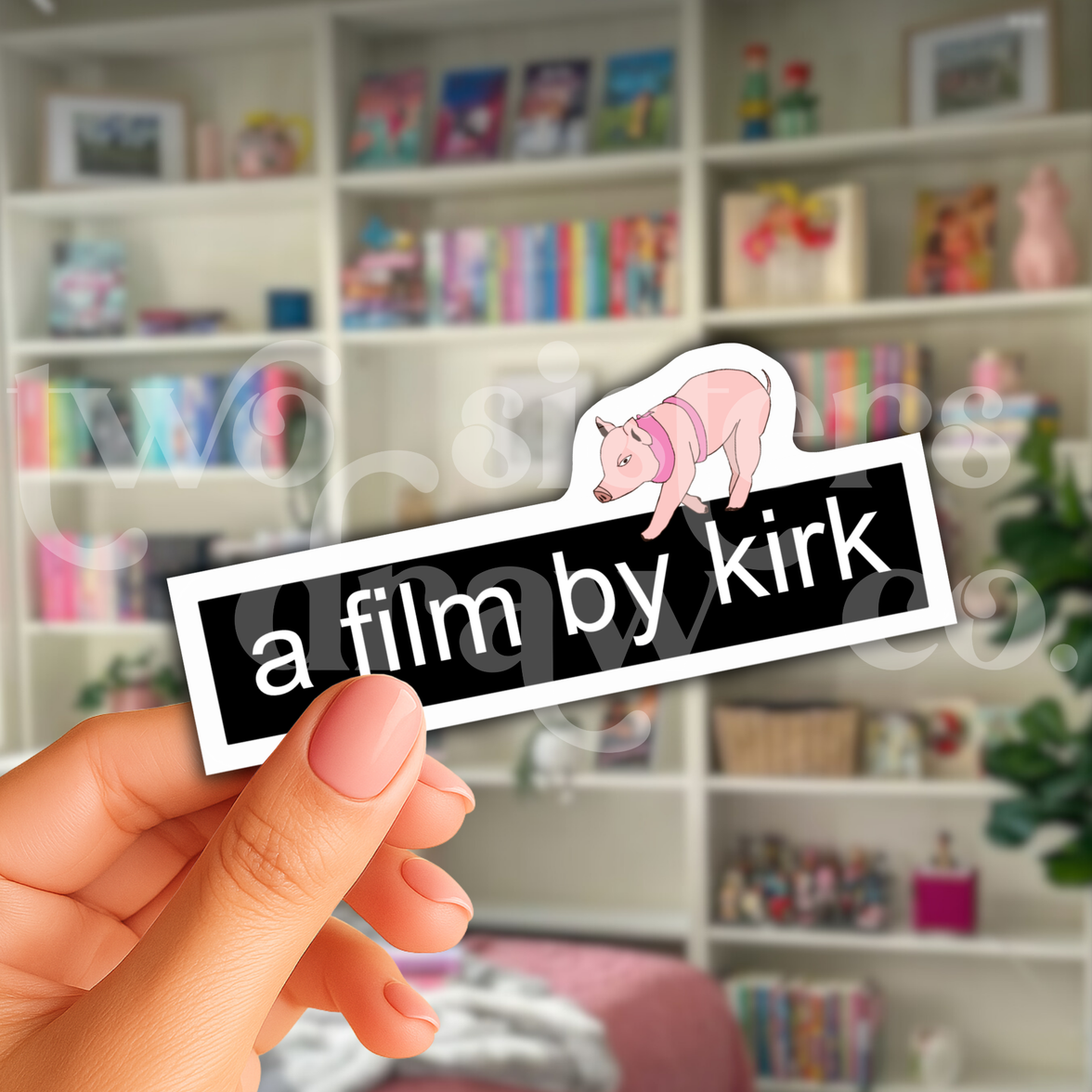 Person holding a Gilmore Girls sticker with the text 'a film by kirk' and a mini pig on top in front of a book case.