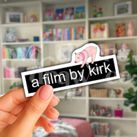 Person holding a Gilmore Girls sticker with the text 'a film by kirk' and a mini pig on top in front of a book case.
