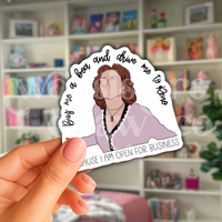 Person holding an illustrated Emily Gilmore sticker with "buy me a boa and drive me to Reno because I am open for business" quote in black script on a white background in front of a bookshelf.