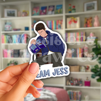 Person holding a sticker of Jess Mariano sitting on a bench reading with 'Team Jess' text in front of a bookshelf.