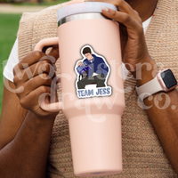 Person holding a tumbler with a sticker of Jess Mariano sitting on a bench reading with 'Team Jess' text.