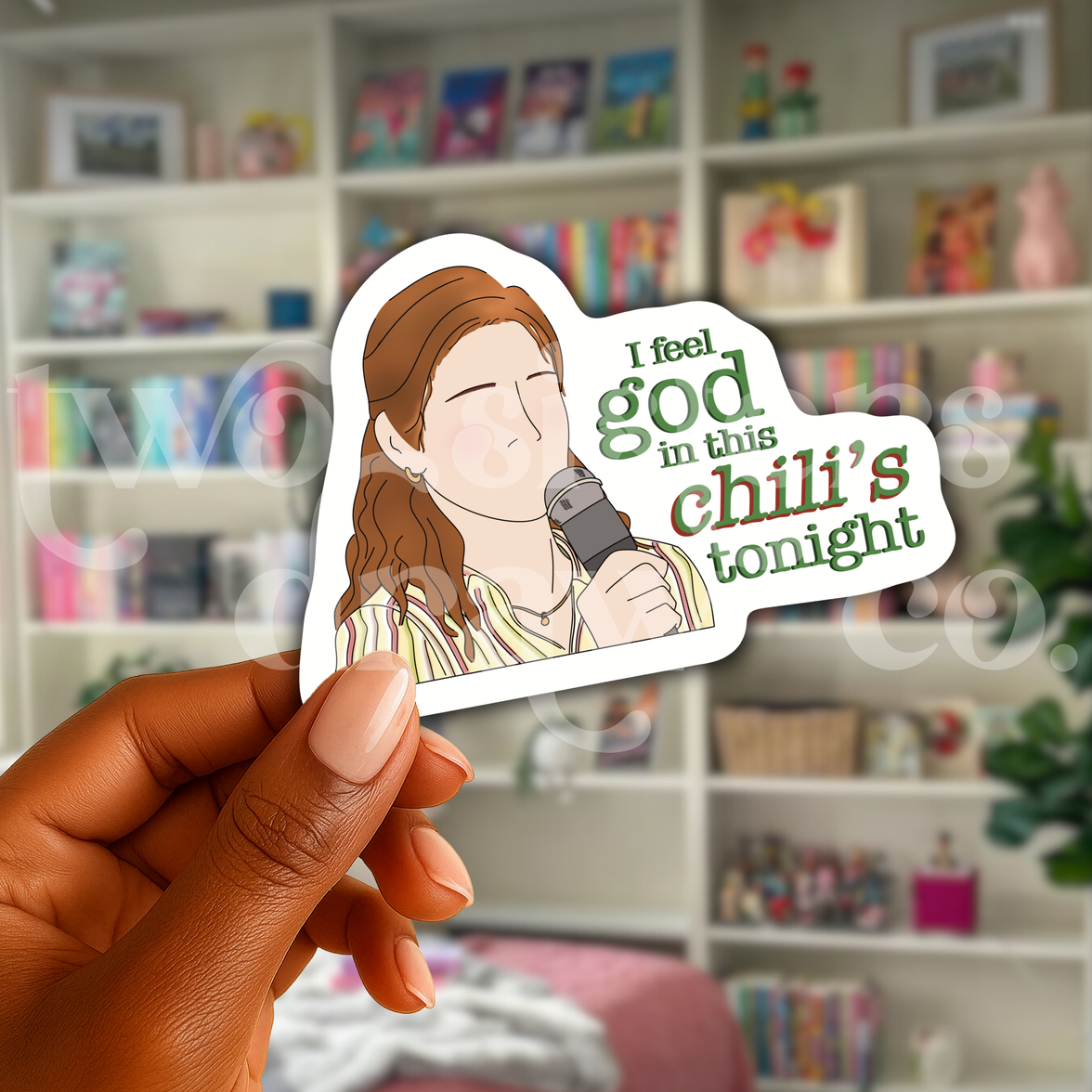 Person holding a hand-drawn sticker of Pam Beesley with a microphone and the text ‘I feel God in this Chili’s tonight’ in front of a book case.