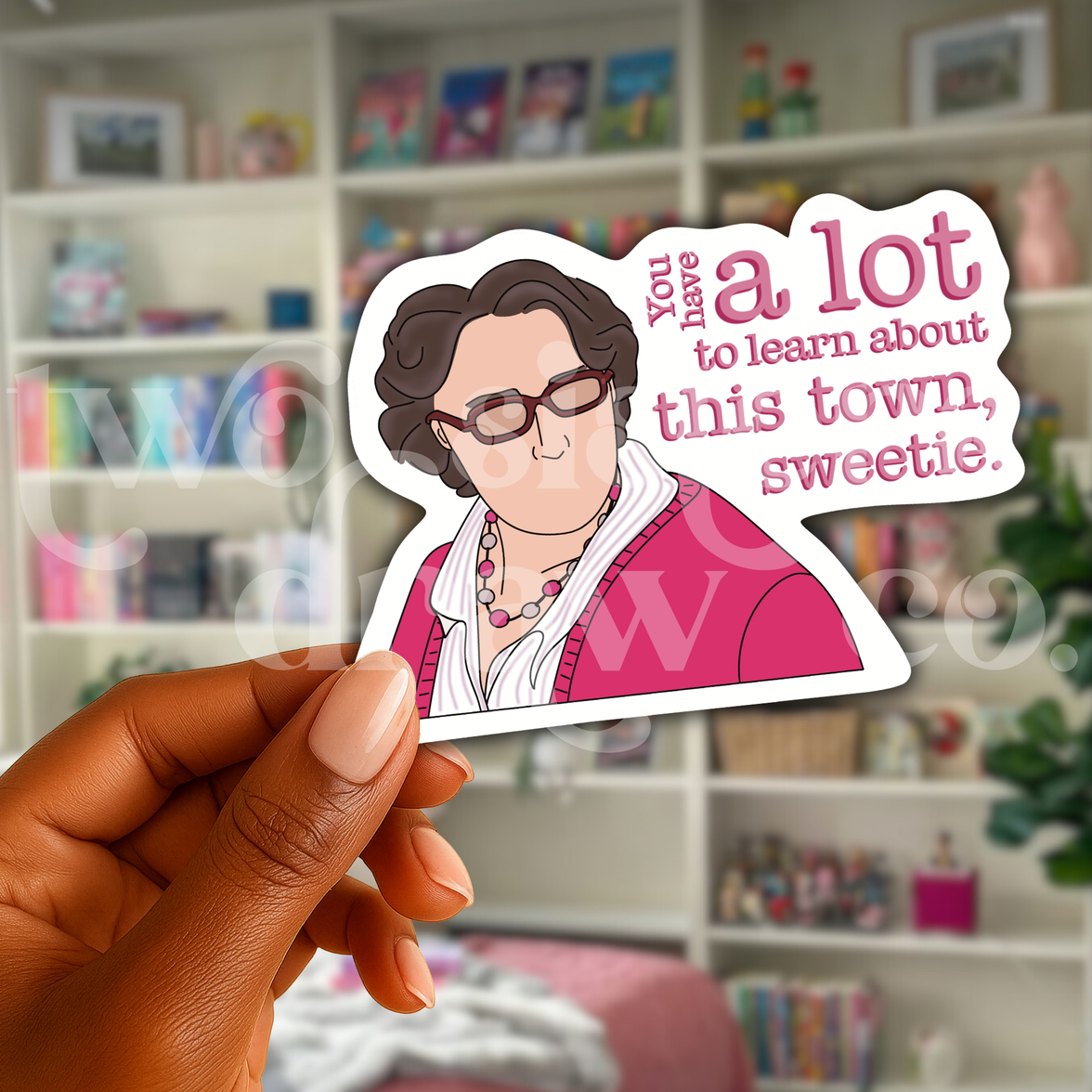 Person holding a hand-drawn sticker of Phyllis Vance from The Office in a pink outfit and glasses with the text ‘you have a lot to learn about this town, sweetie’ in front of a bookcase.
