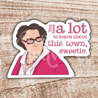 Hand-drawn sticker of Phyllis Vance from The Office in a pink outfit and glasses with the text ‘you have a lot to learn about this town, sweetie’ on a wooden background.