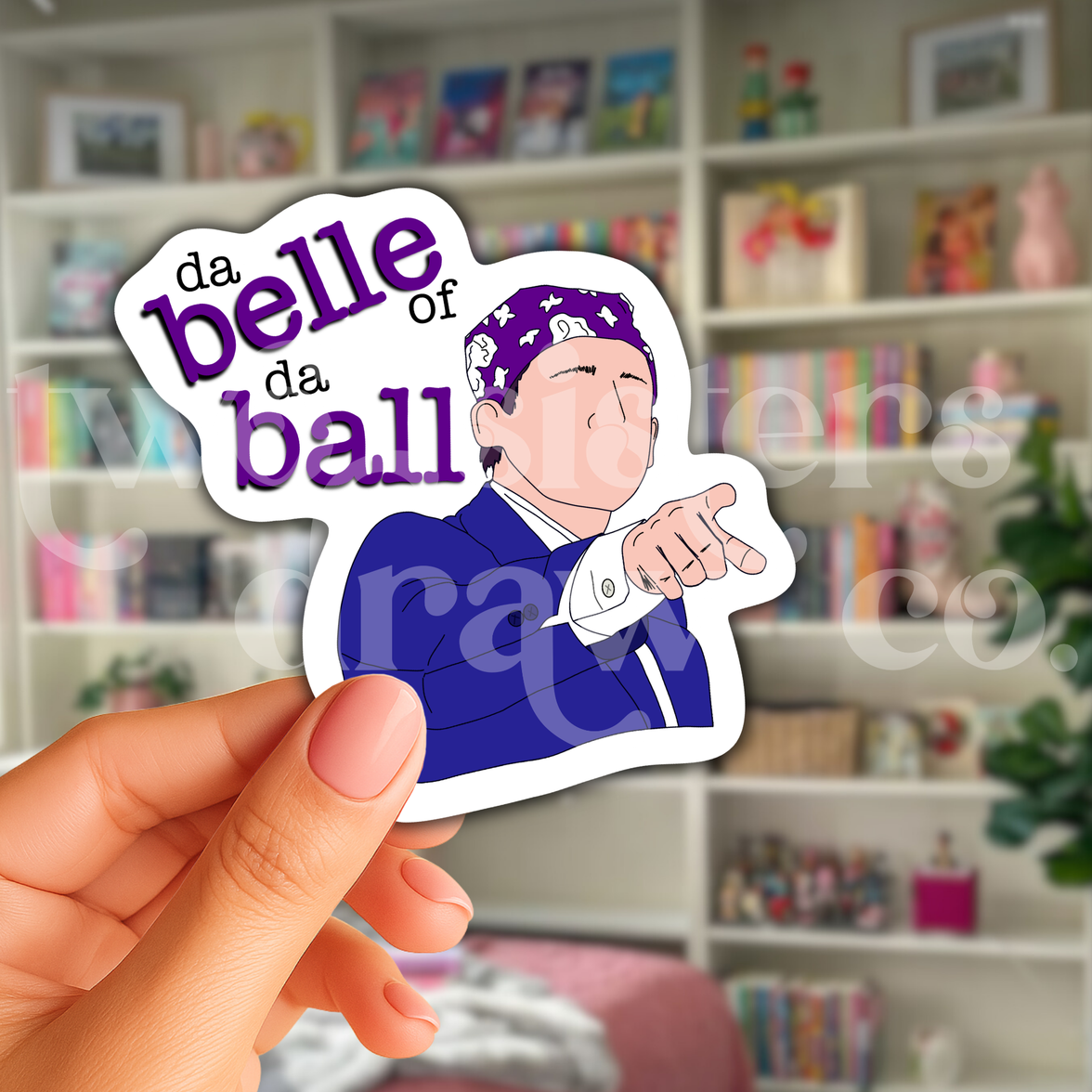 Person holding a hand-drawn sticker of Michael Scott pointing and wearing a purple bandana with the text 'da belle of da ball' in front of a book case.
