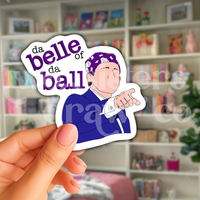 Person holding a hand-drawn sticker of Michael Scott pointing and wearing a purple bandana with the text 'da belle of da ball' in front of a book case.