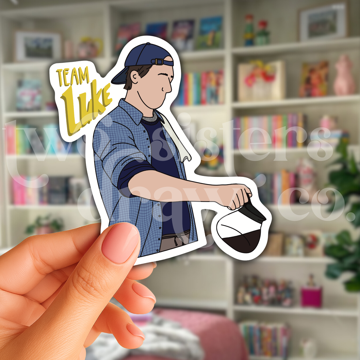 Person holding a hand-drawn sticker of Luke Danes pouring coffee with the test ‘Team Luke’ in front of a book case