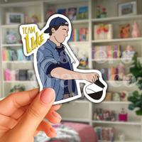 Person holding a hand-drawn sticker of Luke Danes pouring coffee with the test ‘Team Luke’ in front of a book case
