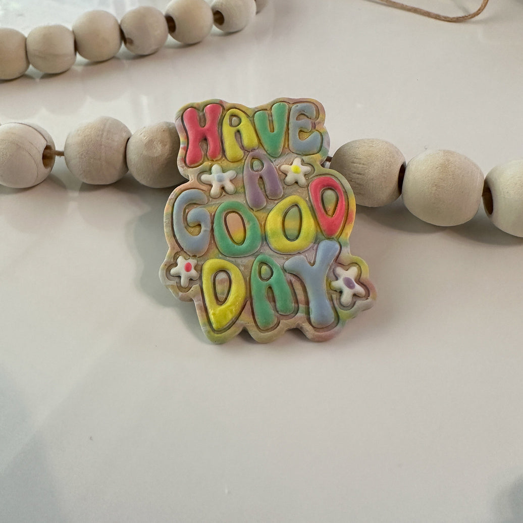 Close-up of handmade, polymer clay magnets featuring 3D bubble lettering  that says 'have a good day' with tiny white flower accents in sherbet colors on a white background with wooden beads.