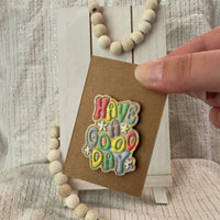 Hand holding a handmade, polymer clay magnets featuring 3D bubble lettering  that says 'have a good day' with tiny white flower accents in sherbet colors on a brown kraft card in front of an easel with wooden beads on a striped cotton background.