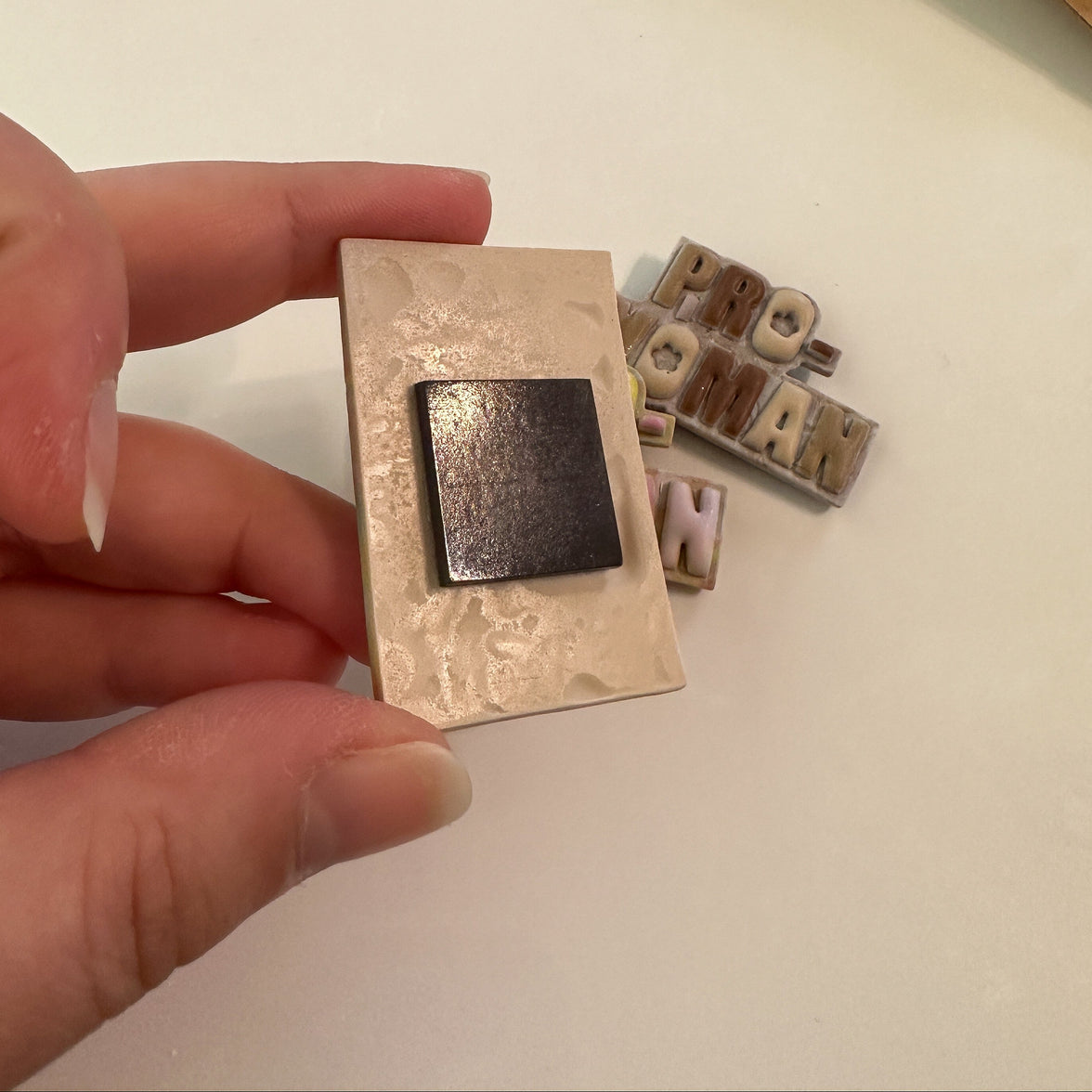 Hand holding a polymer clay magnet showing the adhesive, ferrite magnet secured to the back, with additional magnets behind on a white background.