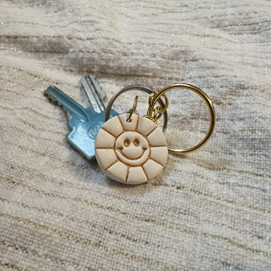 Close-up of a round, white polymer clay keychain featuring a smiley face sun design, and embossed metallic gold liquid clay details with a set of keys on a white cotton background.